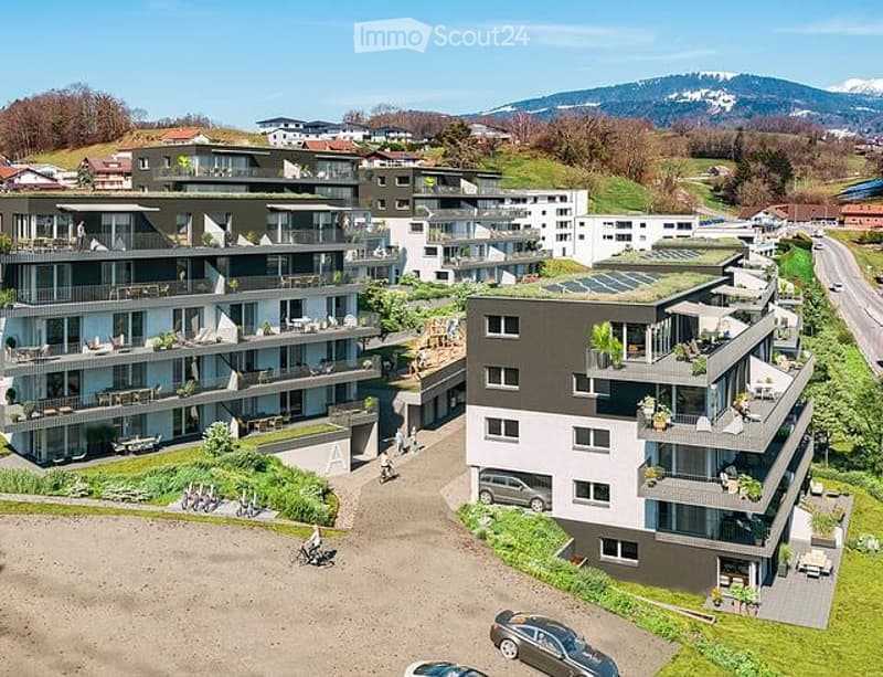 Aerial view of a modern residential complex with multiple apartment buildings, balconies, and surrounding greenery. The buildings feature contemporary architecture with large windows and terraces. There are parking spaces in the foreground and a scenic mo