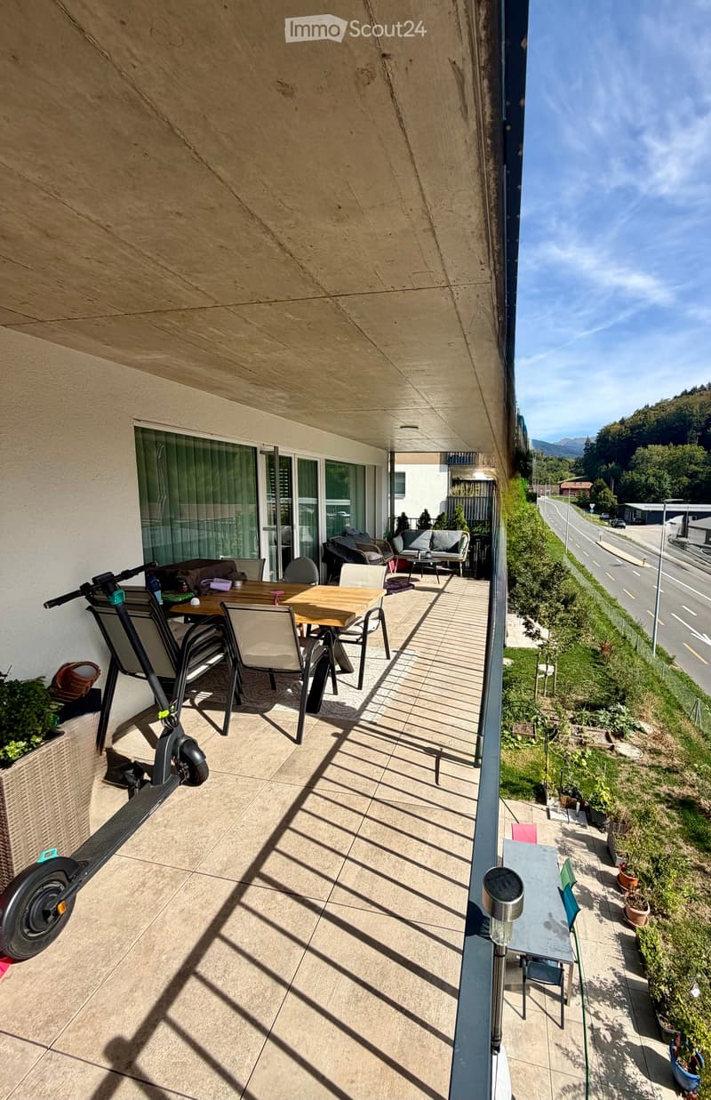 Balcony with a view of a road and greenery, featuring a table, chairs, a sofa, and a scooter.