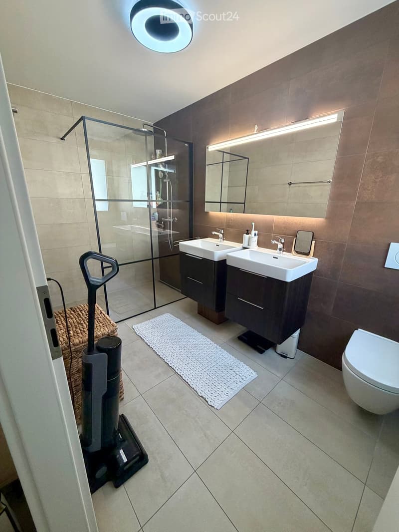 Modern bathroom with double sink vanity, dark wood cabinetry, glass-enclosed shower, and a round LED ceiling light.
