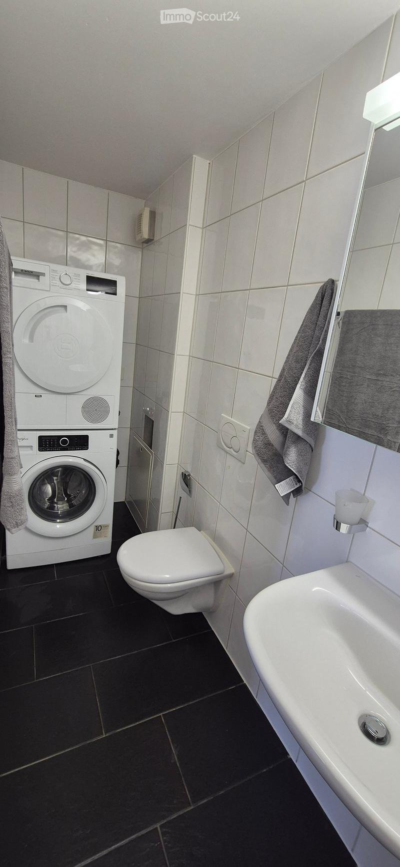 Modern bathroom with white tiles, black floor, toilet, sink, washing machine, and towels.