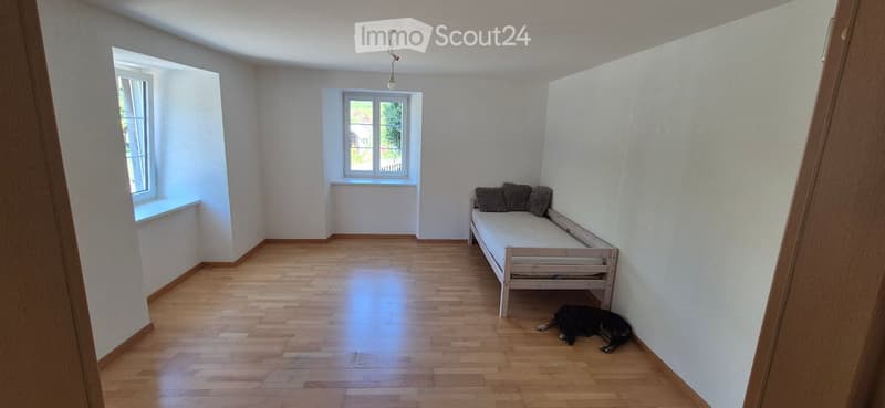 A small, unfurnished bedroom with wooden flooring, white walls, and two windows. A single bed with pillows is positioned against one wall, and a dog is lying on the floor near the bed.