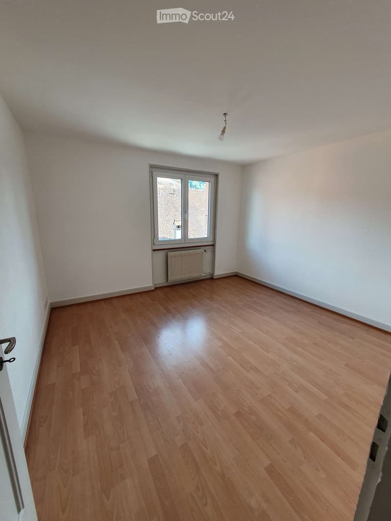 Empty room with wooden flooring, white walls, a window with a view, and a radiator below the window.