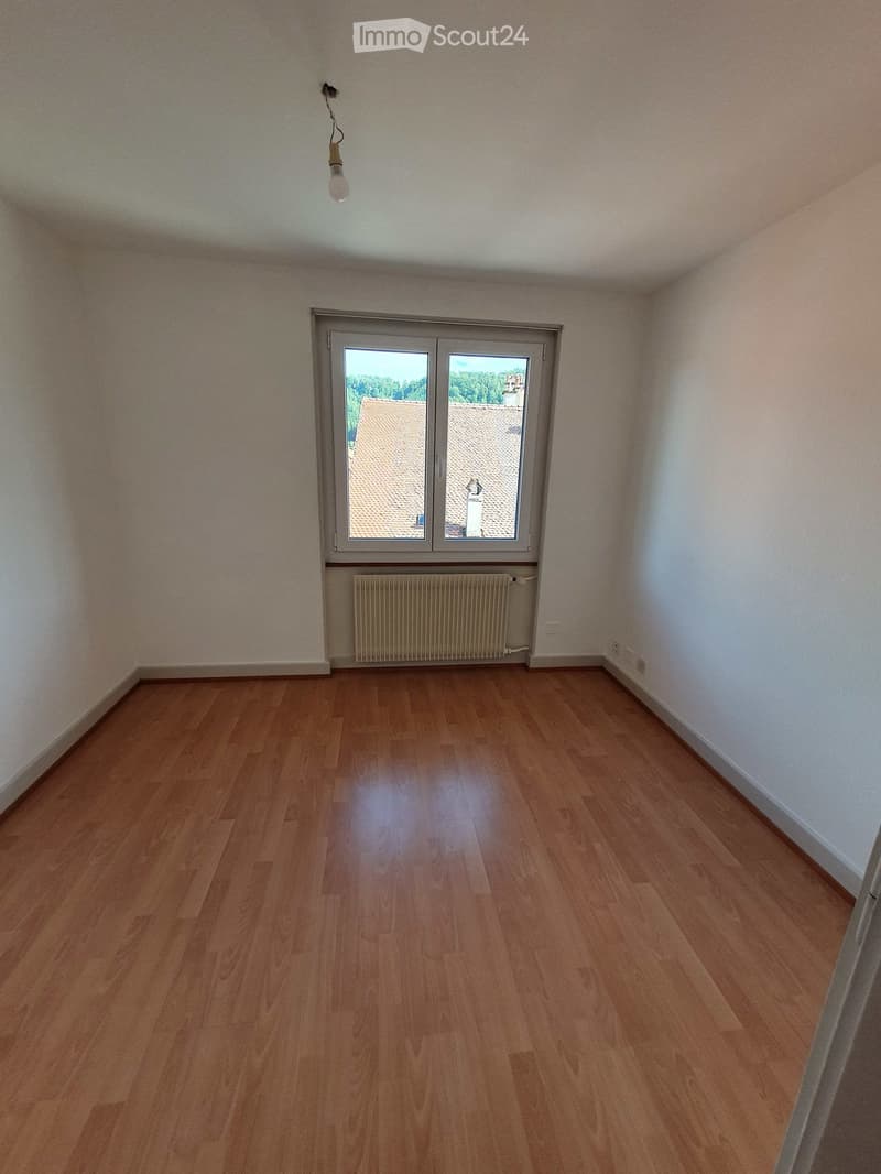 Empty room with wooden flooring, white walls, a window with a view of rooftops, and a radiator below the window.