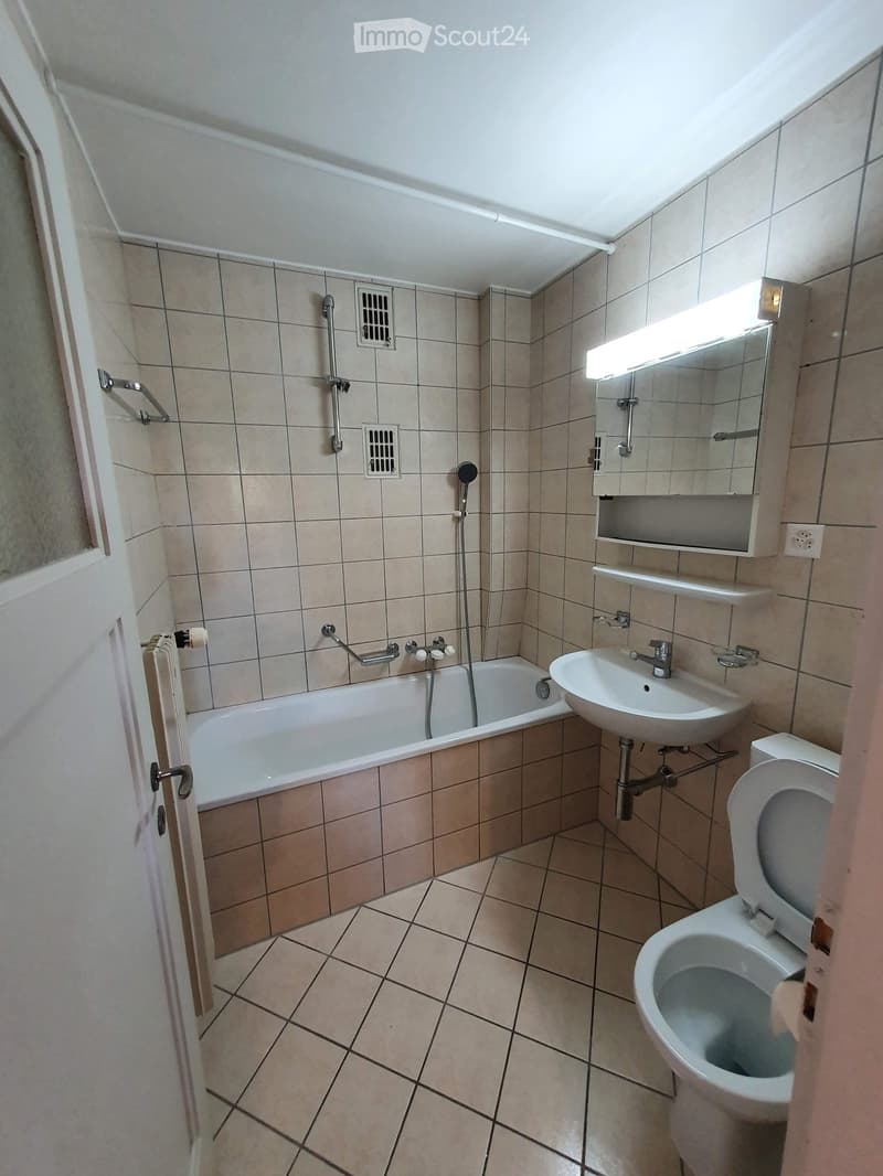 Bathroom with beige tiled walls and floor, featuring a bathtub, sink, toilet, mirror, and showerhead.