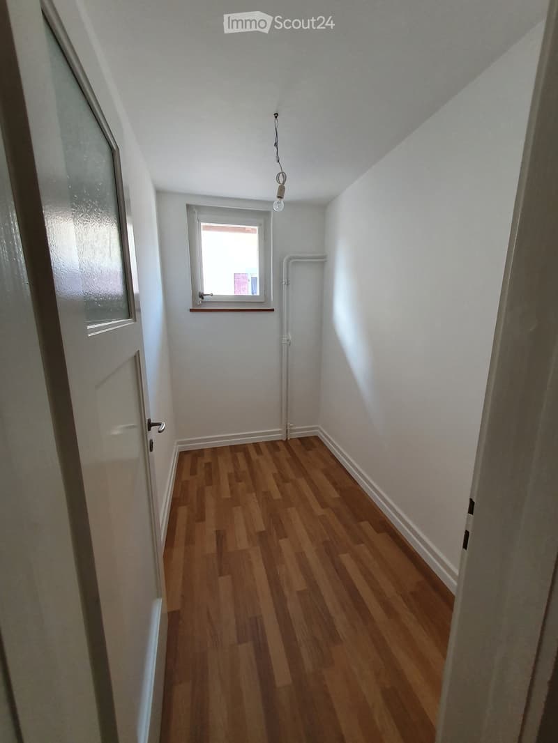 Narrow hallway with wooden flooring, white walls, and a small window at the end. A door is visible on the left side.