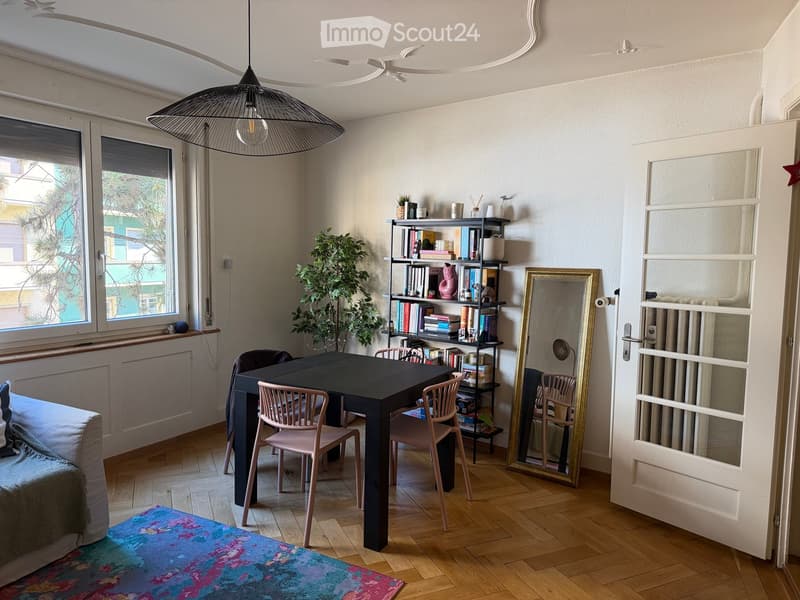 A modern, well-lit room with wooden flooring, a large window, a sofa, a dining table with chairs, a bookshelf, a large mirror, and a hanging pendant light.