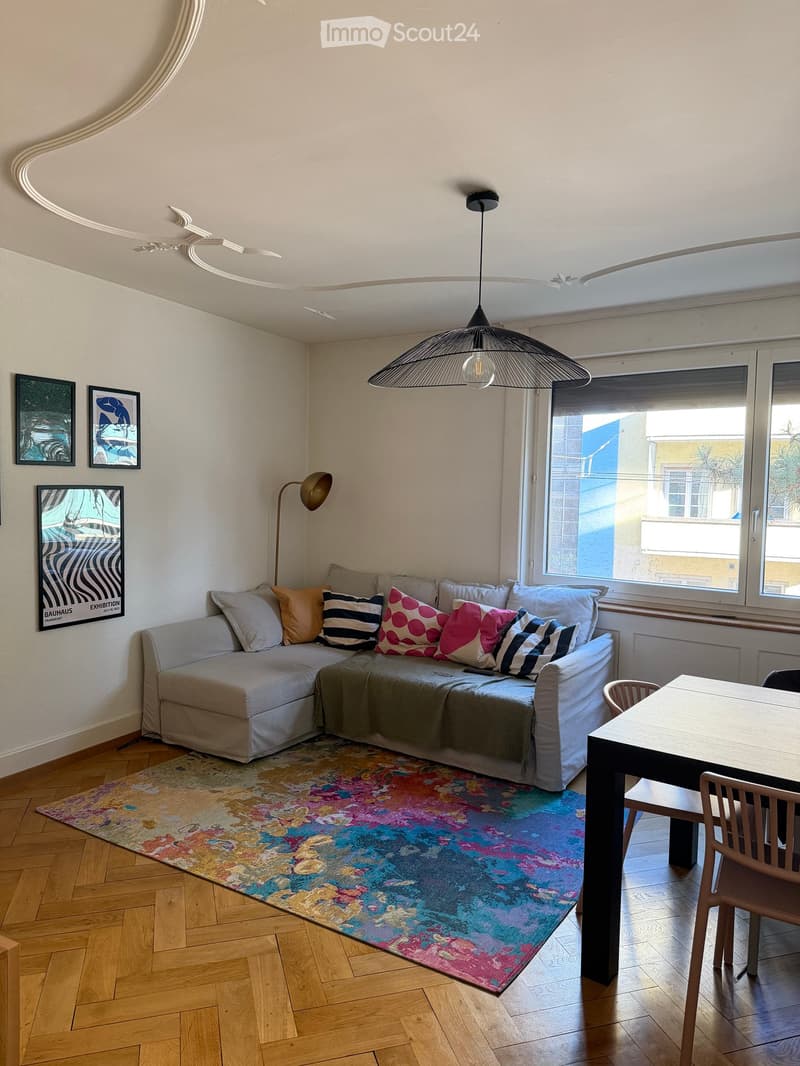 Living room with a grey sectional sofa, colorful throw pillows, a wooden dining table with chairs, a floor lamp, a pendant light, and artwork on the walls.
