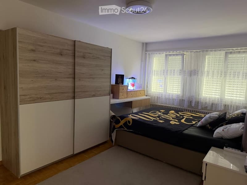 Modern bedroom with a large bed, nightstand, wardrobe, desk, and chair. The room features wooden flooring and large windows with white curtains.