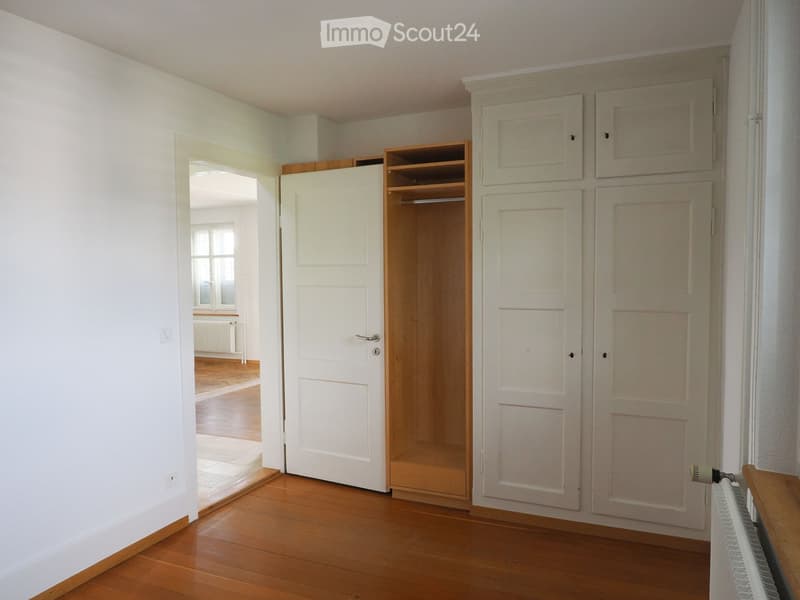 Empty hallway with wooden flooring, white walls, built-in closet with open shelving, and a door leading to another room.