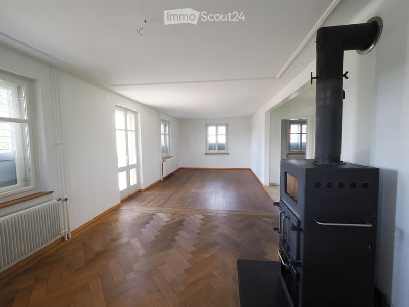 Empty room with wooden flooring, white walls, large windows, and a black wood-burning stove with a chimney.