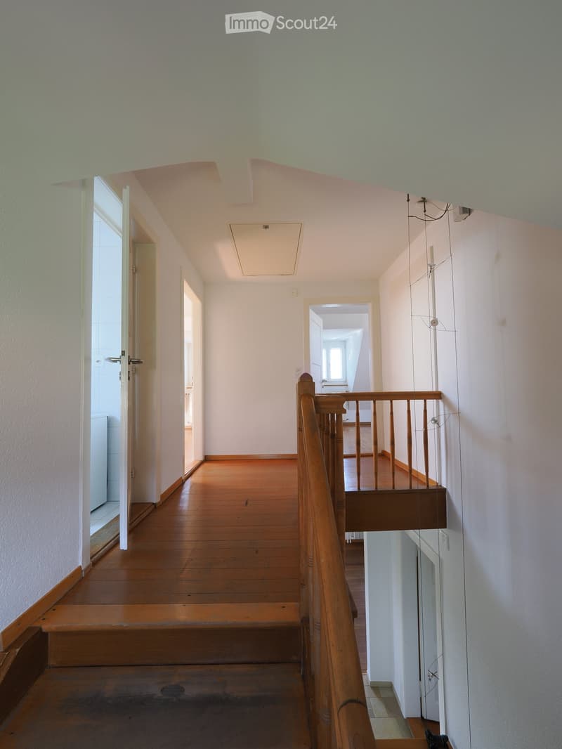 Empty hallway with wooden flooring, white walls, and a wooden staircase leading to an upper level. The hallway has several open doors leading to other rooms.