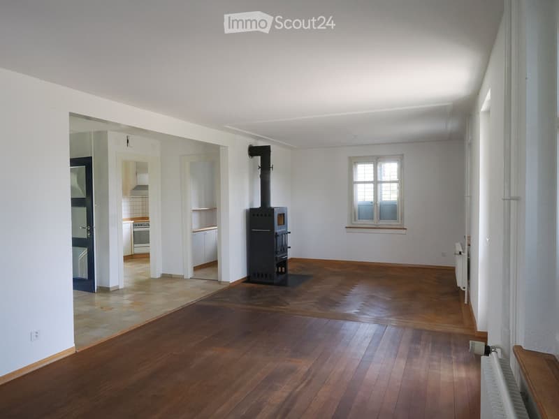 Spacious room with wooden flooring, white walls, and a wood stove in the center. A radiator is visible on the right side, and a doorway leads to a kitchen area with white cabinets and appliances.