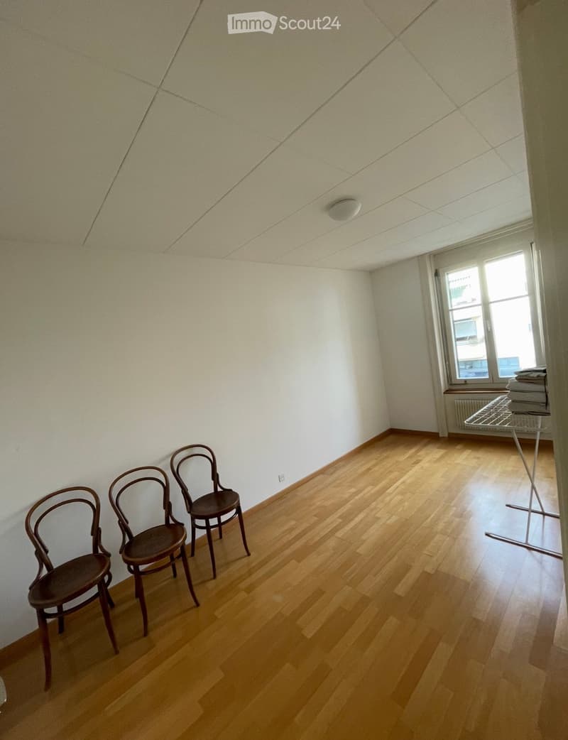 A small, unfurnished room with wooden flooring and white walls, featuring three wooden chairs near the wall and a window with blinds.