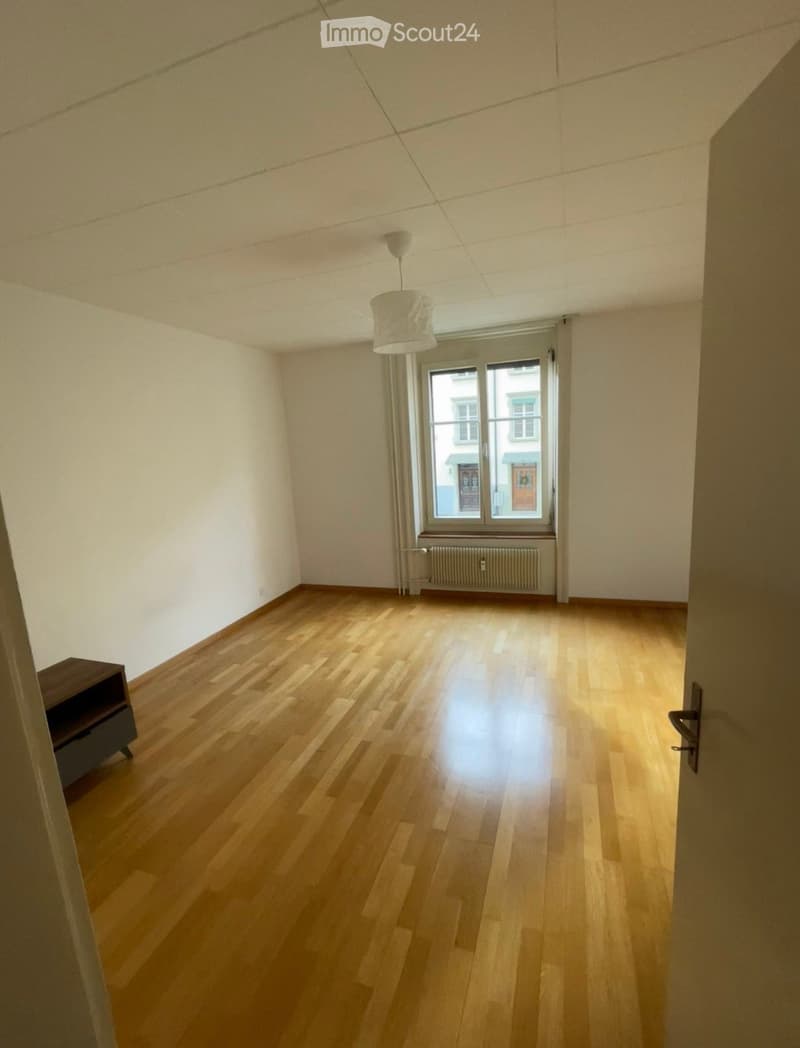 Empty room with wooden flooring, white walls, a window with a radiator below, and a hanging light fixture.