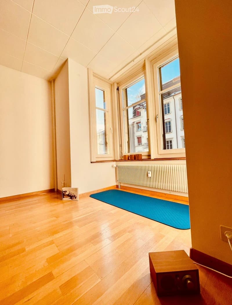 Empty room with wooden flooring, large windows, radiator, yoga mat, speaker, and a book on the floor.