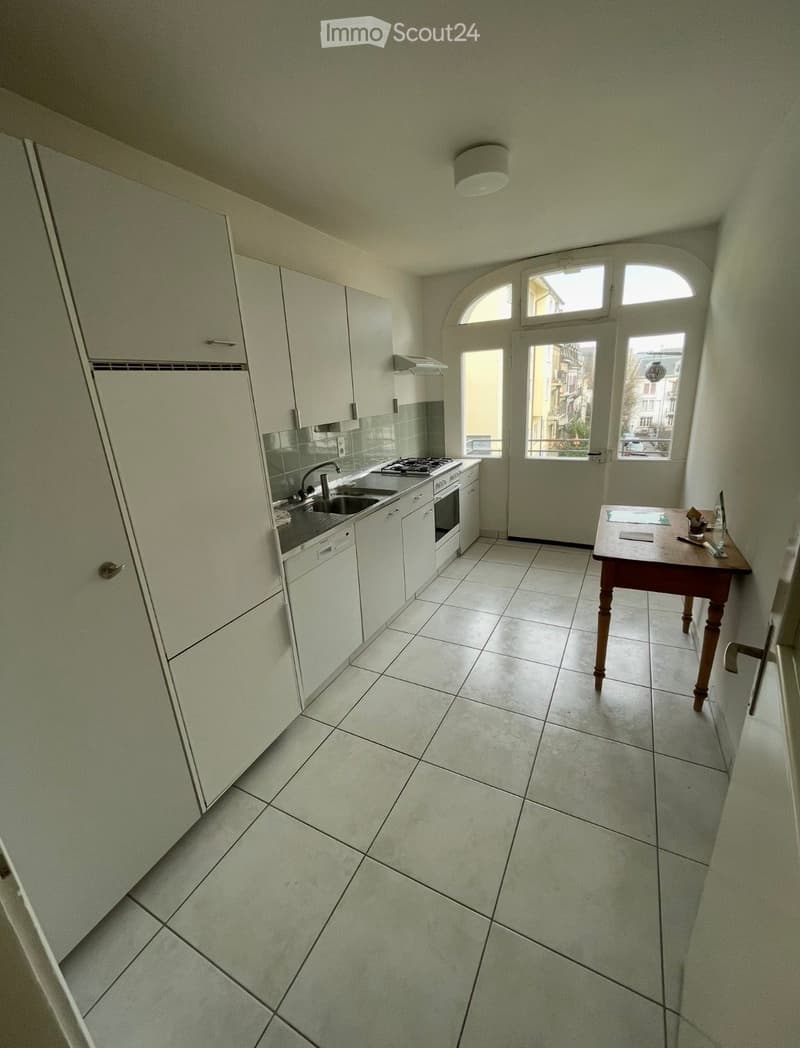 Modern kitchen with white cabinets, tiled floor, sink, oven, dishwasher, and a small wooden table near the window.