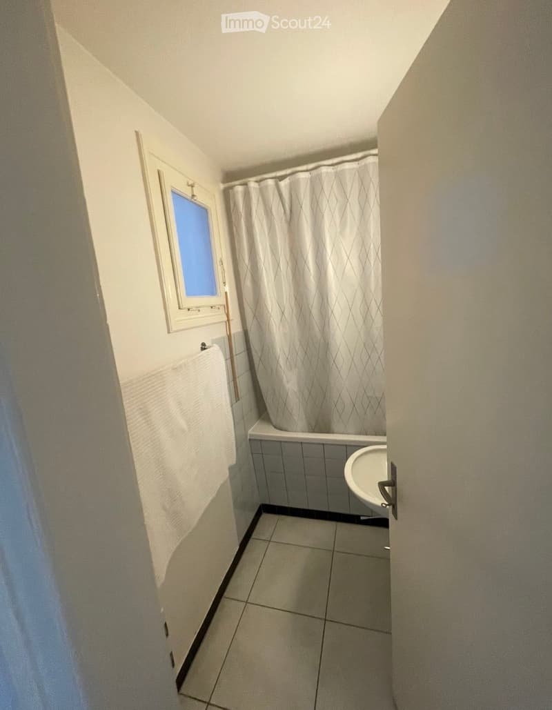 Small bathroom with a bathtub, shower curtain, and sink. Light-colored tiles and a window provide natural light.