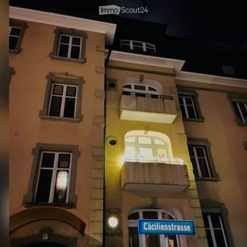 Multi-story residential building with illuminated balconies and windows, featuring a street sign reading "Cäcilienstrasse".