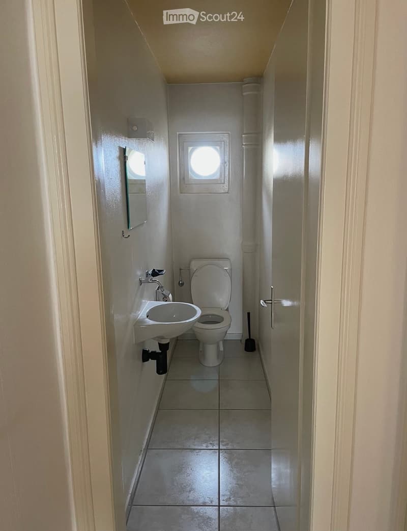 Small bathroom with a toilet, sink, mirror, and a small window above the toilet. The walls are tiled, and the floor is made of large square tiles.