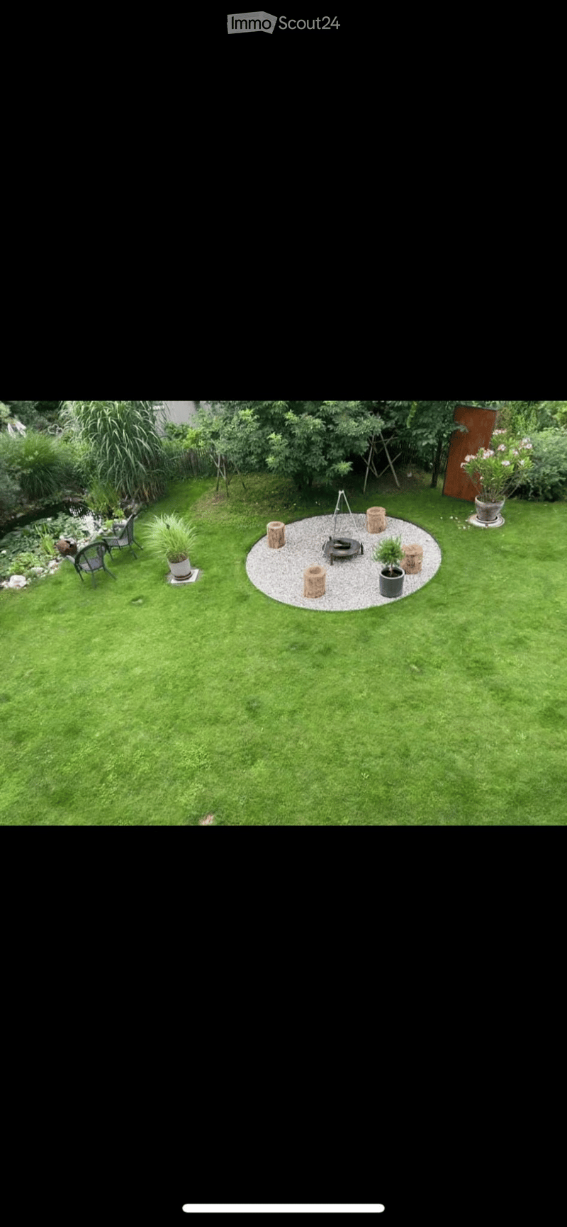 Well-maintained garden with a circular gravel fire pit surrounded by wooden logs, potted plants, and a small seating area with chairs and a table.