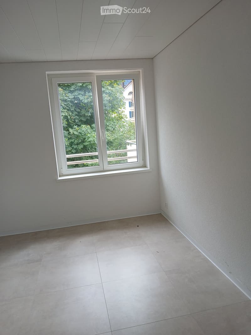 Empty room with light grey walls, white tiled floor, and a large window with white frame showing greenery outside.