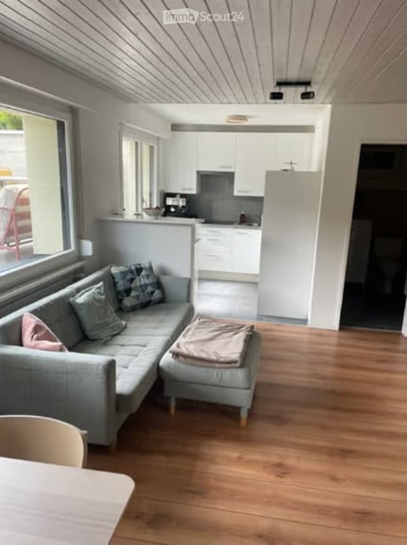 Open-plan living and dining area with a grey sectional sofa, ottoman, and a chair. The kitchen features white cabinets and a refrigerator. The space has wooden flooring and large windows.