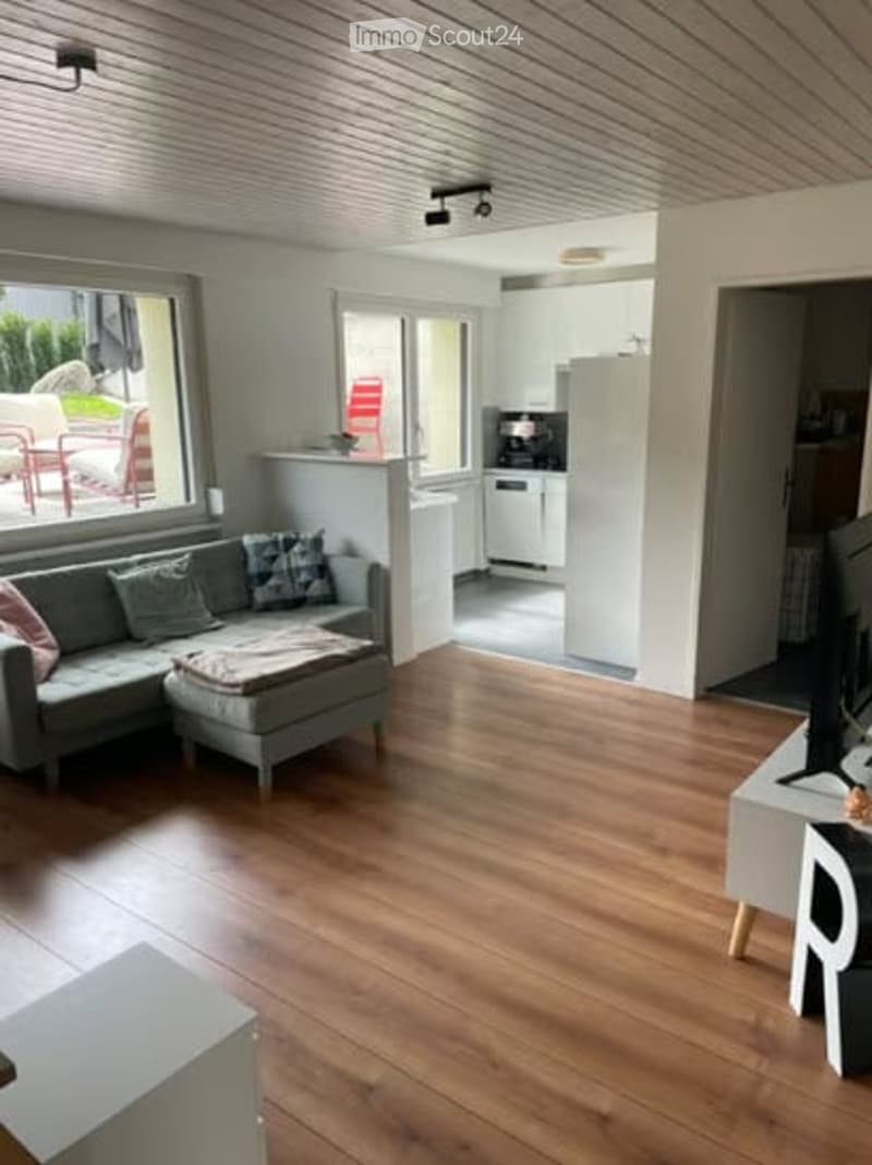 Bright living room with wooden flooring, grey sectional sofa, ottoman, small table, and television. The room has large windows and an open kitchen area visible in the background.