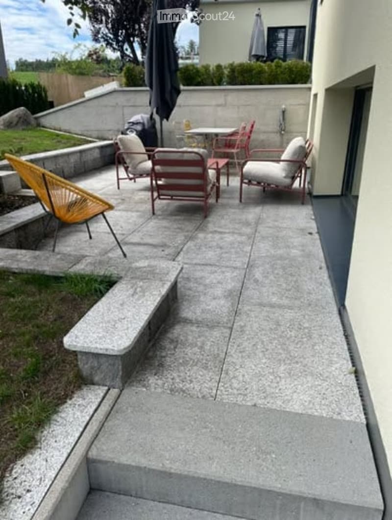 Outdoor patio area with concrete flooring, several chairs and tables, umbrella, and steps leading up to a raised section.