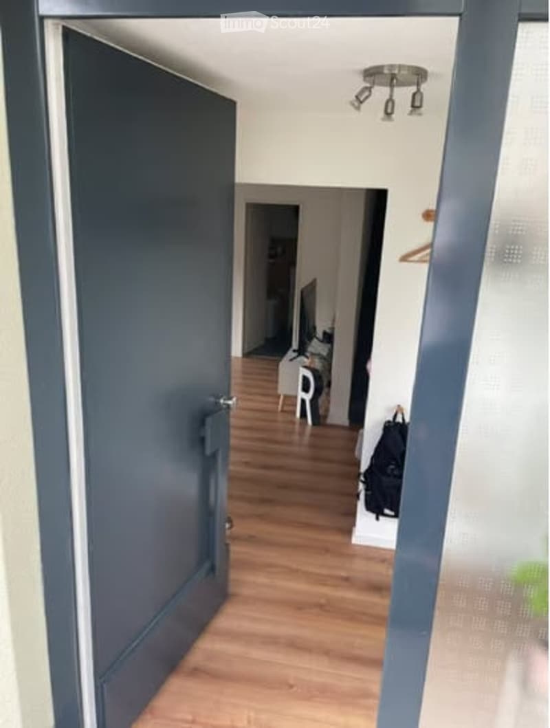 A narrow hallway with wooden flooring, a closed dark blue door on the left, and an open doorway leading to another room with visible furniture.