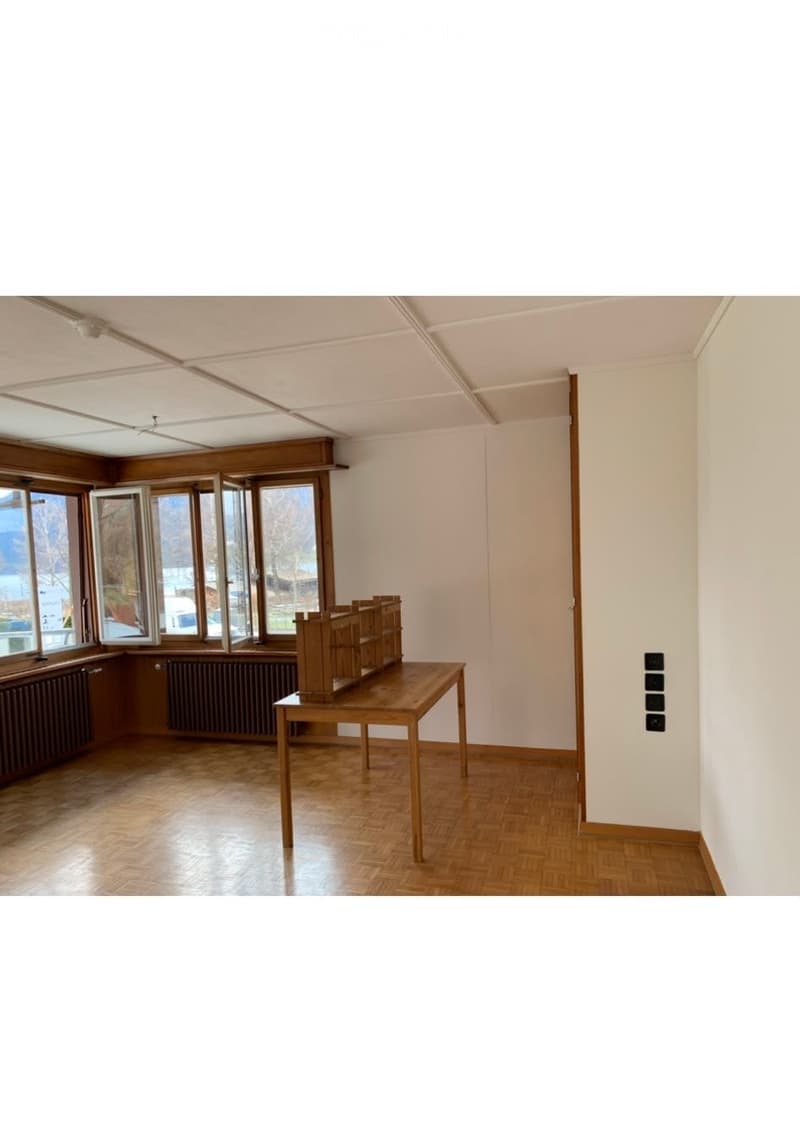 Bright room with wooden flooring, large windows, a wooden table with shelves, and white walls.