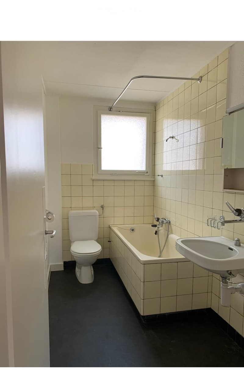 Bathroom with white tiles, a toilet, a bathtub, a sink, and a showerhead. The room has a window and a dark floor.