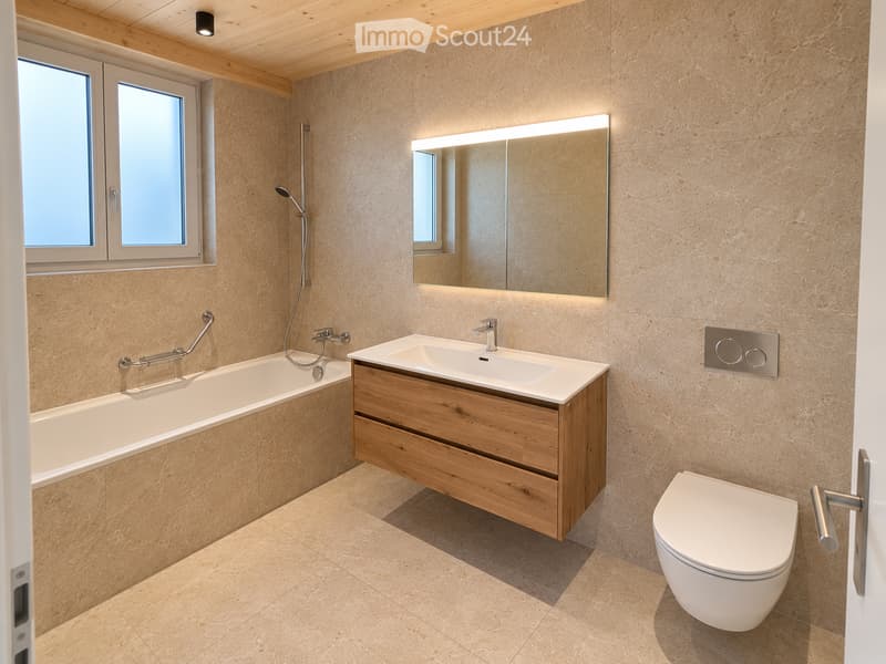Modern bathroom with beige tiles, a wooden sink cabinet, a bathtub with grab bars, a toilet, and a large illuminated mirror above the sink.