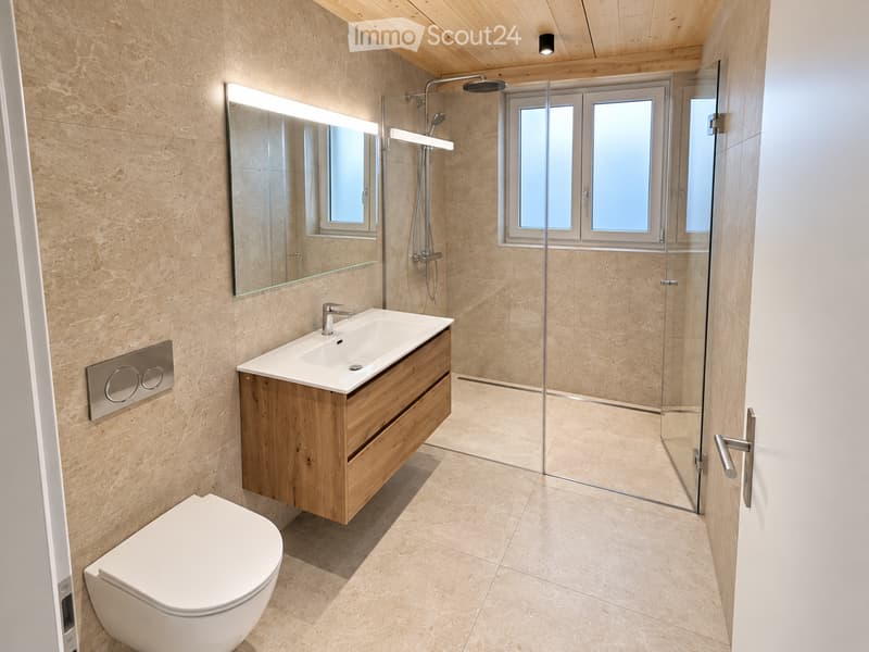 Modern bathroom with a wooden vanity, white sink, toilet, large mirror, and a glass-enclosed shower area with a window.