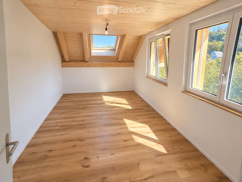 Empty room with wooden flooring, white walls, and large windows offering a scenic view of trees and mountains. The room features a skylight and a modern, minimalist design.