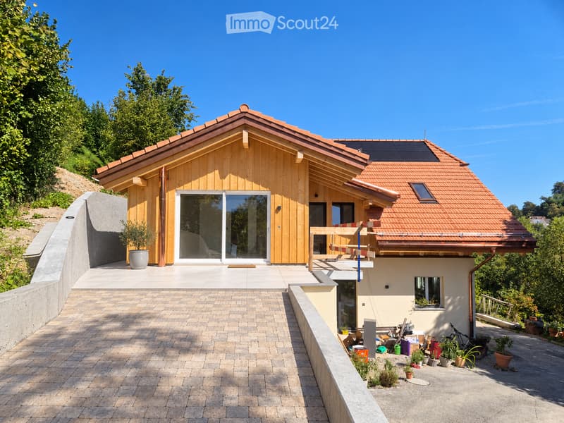 Modern two-story house with wooden facade, red-tiled roof, large windows, and solar panels on the roof. The house is situated on a raised ground floor with a paved pathway leading to the entrance.