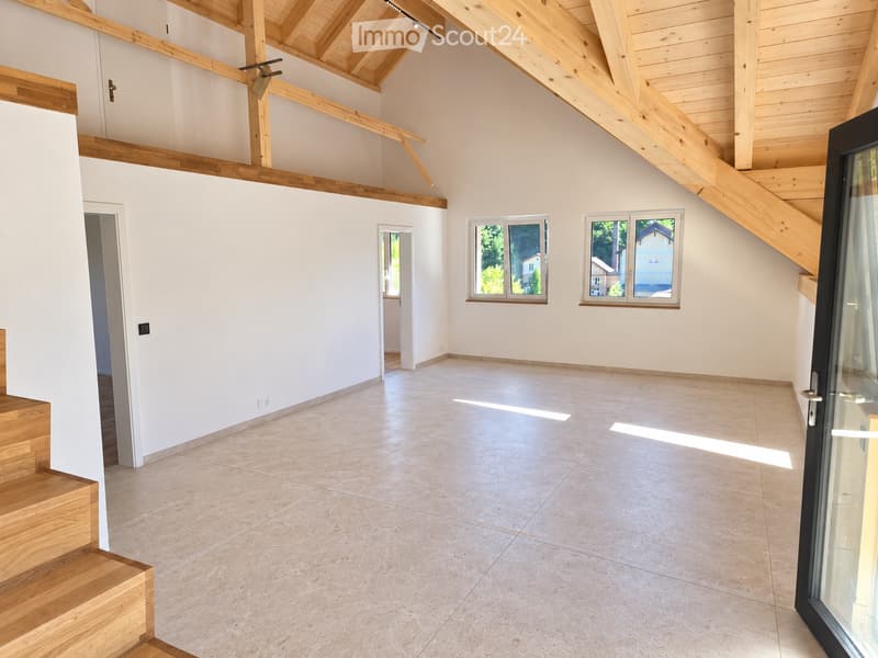 Bright, empty room with white walls, wooden beams, tiled floor, and large windows letting in natural light. Stairs lead upwards.