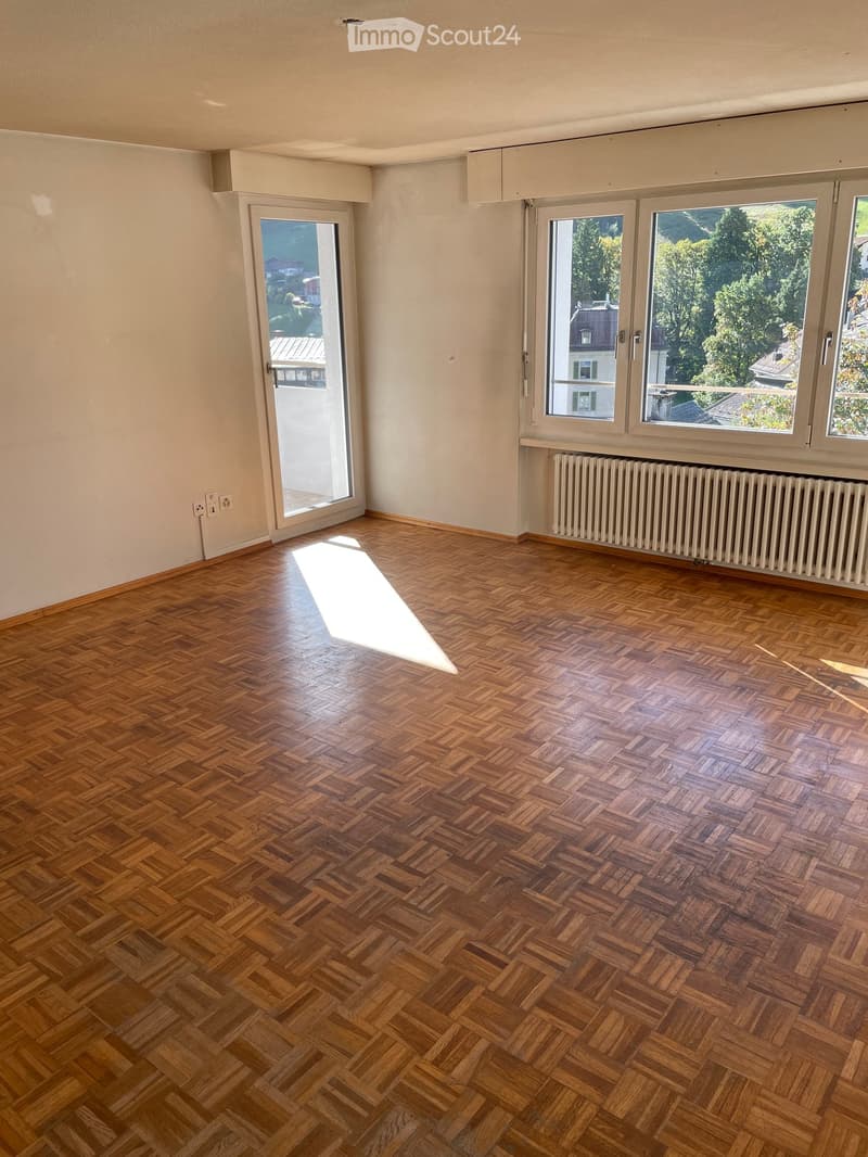 Empty room with wooden herringbone flooring, large windows, sliding glass door, radiator, and visible electrical outlets.