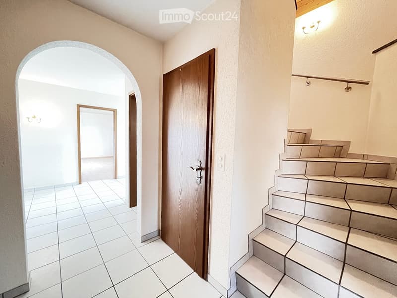 Hallway with tiled floor, wooden door, staircase leading upwards, and arched doorway to another room.