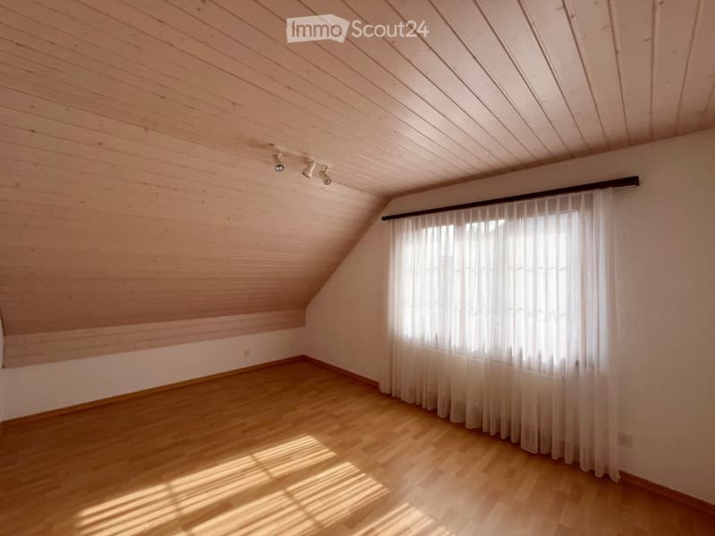 Empty attic room with wooden walls, wooden floor, large window with white curtains, and recessed lighting.