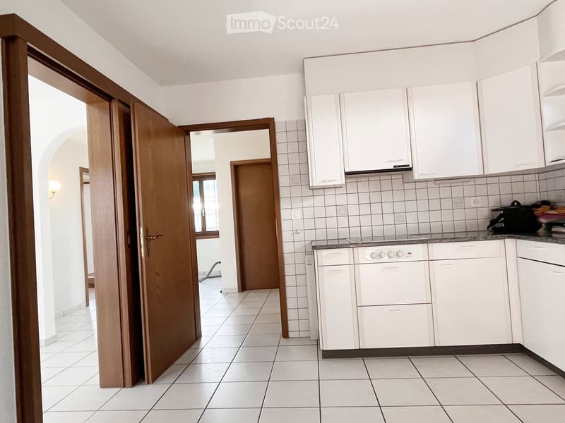 Bright kitchen with white tiled walls and floor, white cabinets, and a stove. An open wooden door leads to another room.