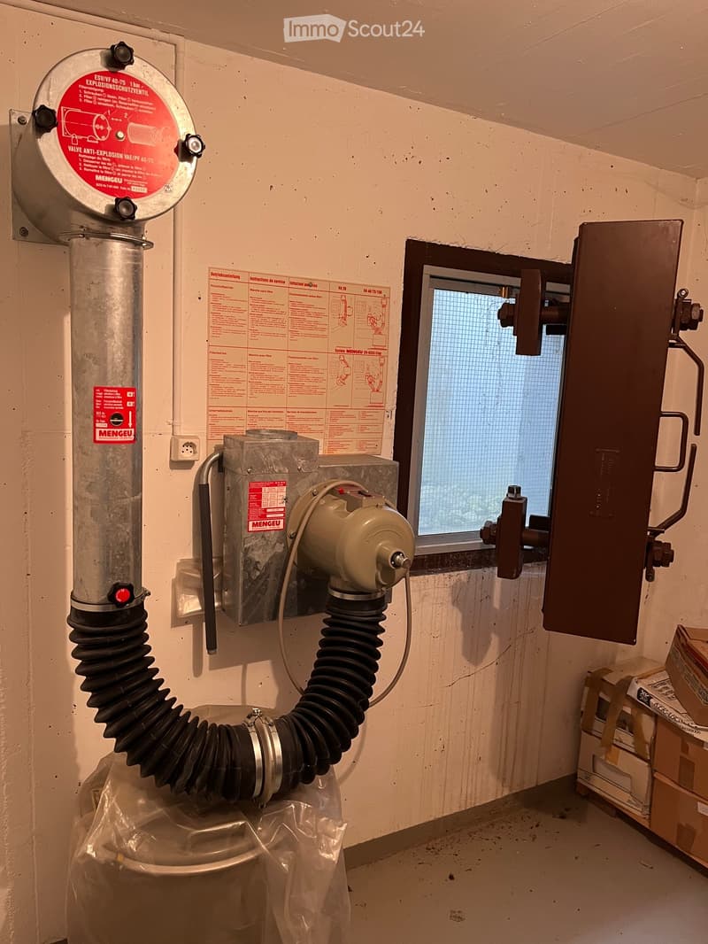 An unfurnished room with a ventilation system, including a large metal pipe, a window with a grid, and various mechanical components attached to the wall.
