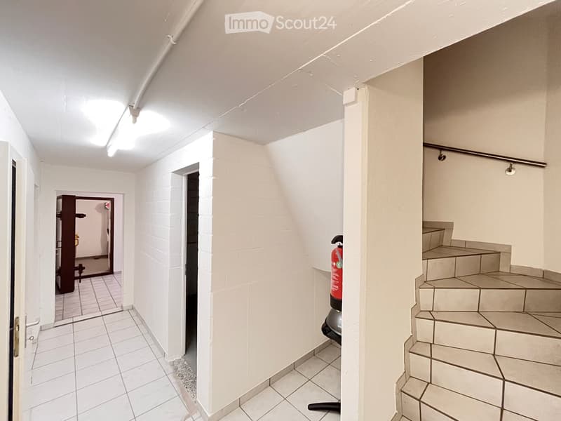 A narrow, white-walled hallway with tiled flooring, leading to a staircase and an open doorway. A fire extinguisher is placed near the staircase, and a small shelf is visible in the adjacent room.