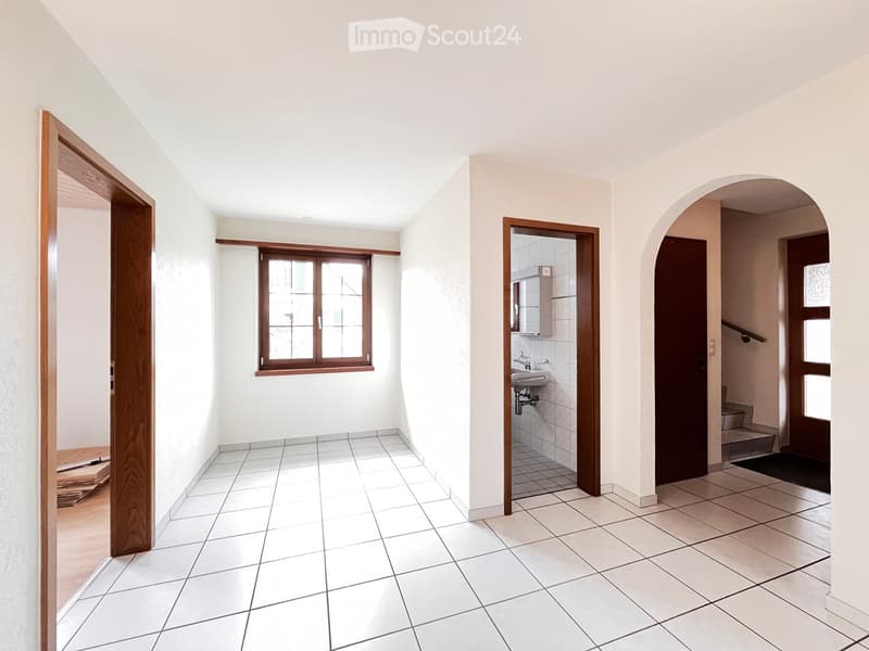 Empty hallway with tiled floor, white walls, and wooden doors leading to a bathroom and staircase area.