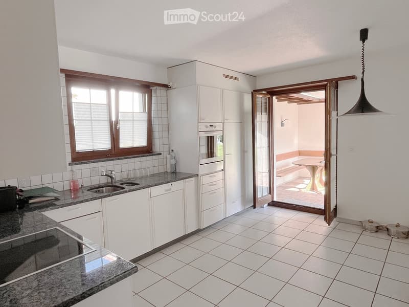 Modern kitchen with white cabinets, granite countertops, stainless steel sink and faucet, built-in oven and dishwasher, and a window with blinds. An open doorway leads to a patio area with a table and seating.