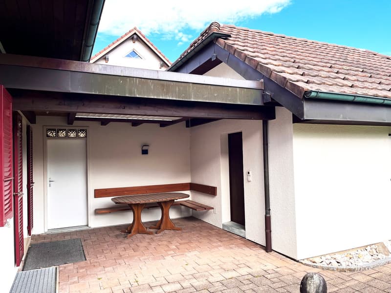 Outdoor seating area with a wooden table and benches under a covered patio attached to a house.