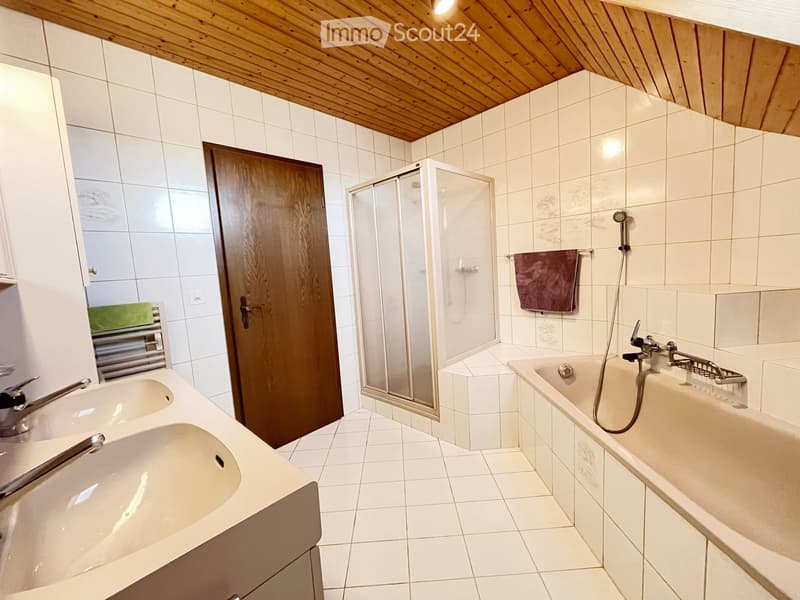 Modern bathroom with white tiled walls and floor, a wooden slanted ceiling, a bathtub with a handheld shower, a double sink vanity, a glass-enclosed shower stall, and a towel hanging on a rack.