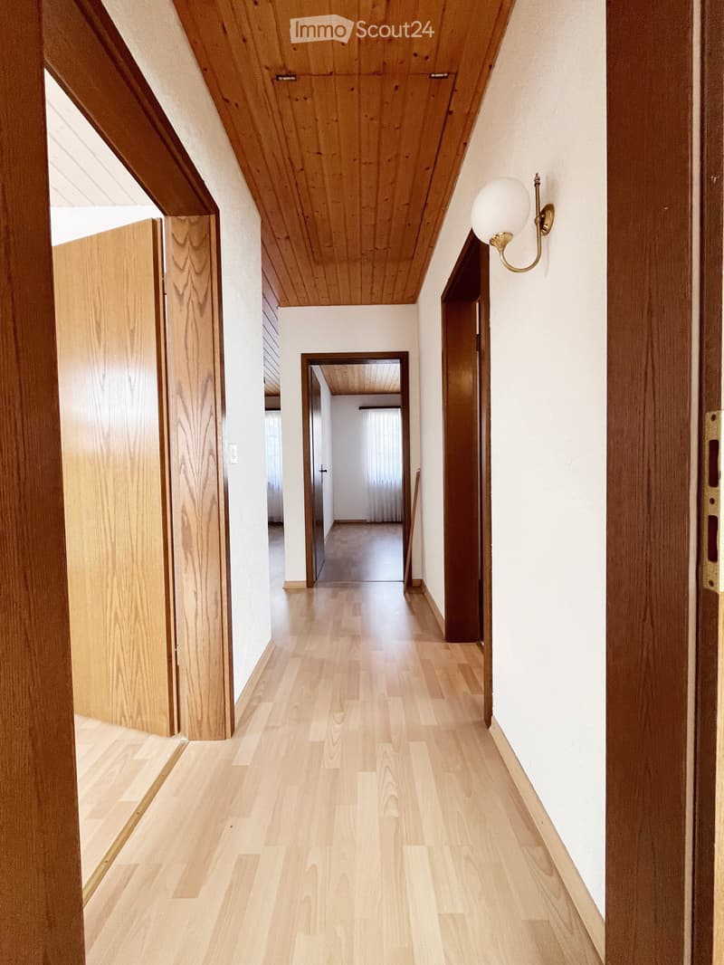A modern hallway with light wooden flooring, white walls, and wooden doors. The ceiling has a slanted, wooden design, and a wall-mounted light fixture is visible on the right side.
