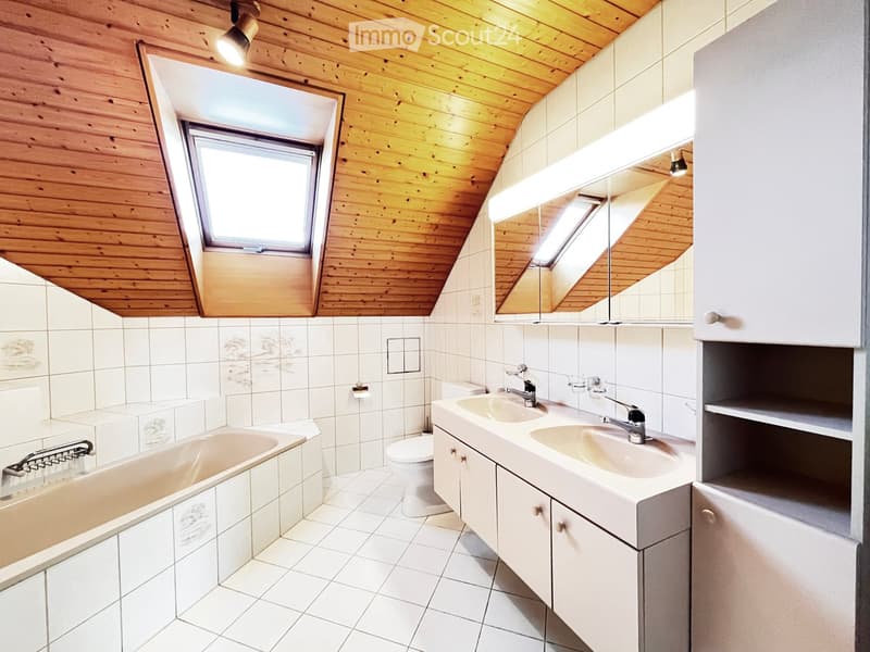 Bathroom with wooden ceiling, skylight windows, white tiled walls and floor, bathtub, toilet, double sink, and mirrored cabinet.