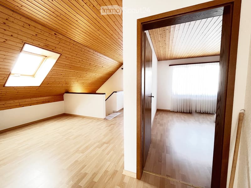 Empty room with wooden flooring and slanted wooden ceiling, featuring a skylight window and a doorway leading to another room with white walls and wooden flooring.