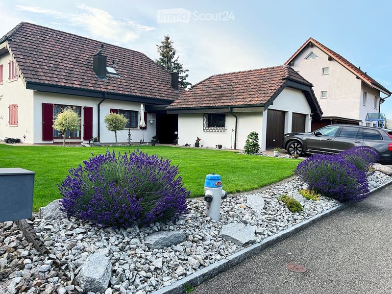 Two-story house with red-tiled roof, white walls, and red shutters. The front yard features a well-maintained lawn, lavender bushes, and a fire hydrant.